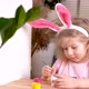 a Little Blonde Girl is Decorating an Easter Egg and Smiling at Home at the Table