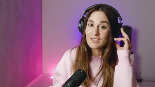 Happy Young Woman Taking Off Headphones Looking at Camera and Talking Smiling While Sitting at Desk alt
