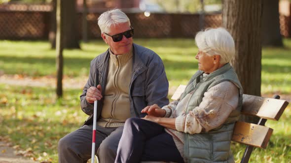 Blind Elderly Man Sitting with His Wife on the Bench in the Park and Holding Blind Cane alt
