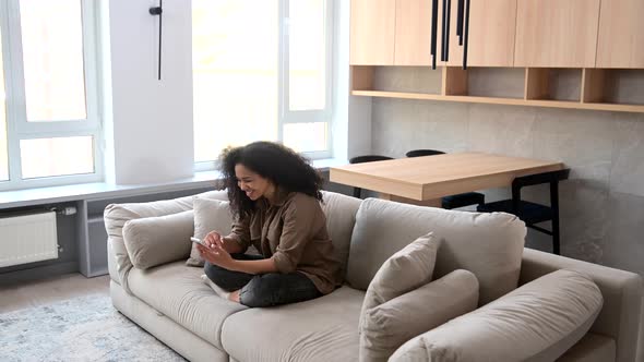 Side View at Young Multiracial Woman Using Trendy Smartphone Sitting on the Sofa at Home