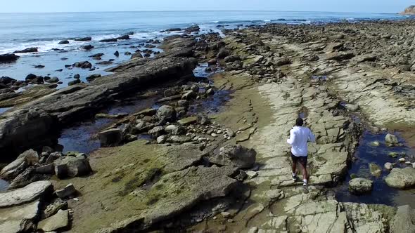Tracking shot of a young man running on a rocky ocean beach shoreline alt