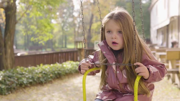 Cute Little Caucasian Girl Talks Then Looks Around As She Sits on a Swing Ride alt