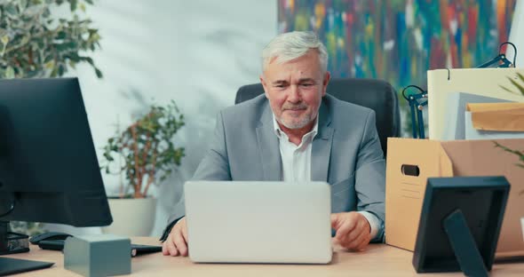 Man in Suit with Gray Hair Leads Remote Conversation From Office Sitting at Desk in Front of Laptop alt