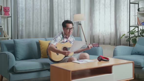 Asian Man Composer With Note Book And Looking At Paper While Playing Guitar At Home alt
