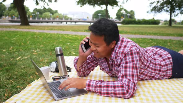 Happy Aisan man lying, working on laptop and having a call at the park alt