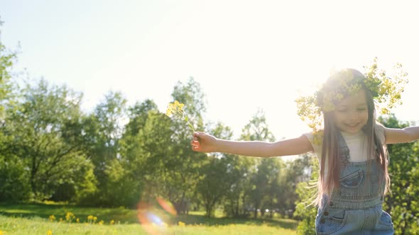 Cute Little Girl in a Flower Wreath on Her Head Jumps and Rejoices Among Dandelions in a Spring alt