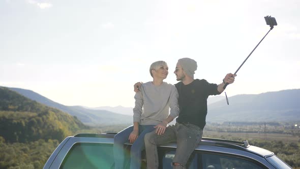 Happy Couple Sitting on Roof of Car and Possing on the Smart Phone Camera in the Mountain alt
