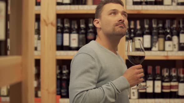 Handsome Man Standing with Glass of Red Wine in Liquor Store. Woman in the Background alt