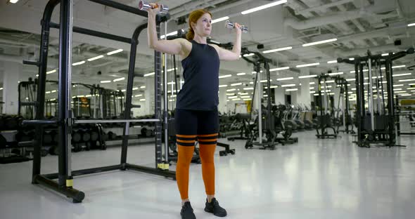 Athletic Girl in the Gym Doing Standing Exercises with Dumbbells on Her Arms and Shoulders alt