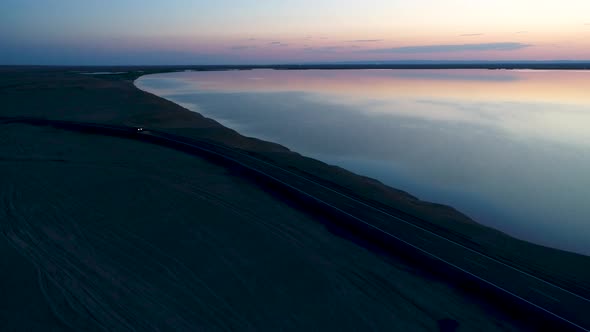 Aerial View of Car Driving on an Empty Road in the Desert Next To the Lake During Sunset alt