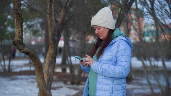 Brunette Woman Is Browsing In Her Mobile Phone Standing In Park Early Spring