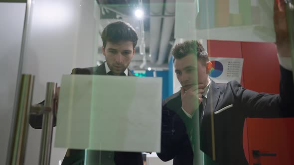 Two Thoughtful Men Analyzing Strategy Looking at Paperwork Standing Behind Glass Door in Office alt