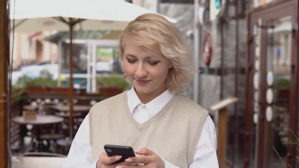 Young Blonde Woman with Elegant Manicure in a Beige Vest and White Blouse Swipes in a Cell Phone alt