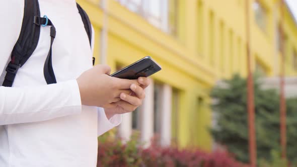 A Caucasian Teenage Boy Looks at a Smartphone  Closeup on the Phone  a School in the Background