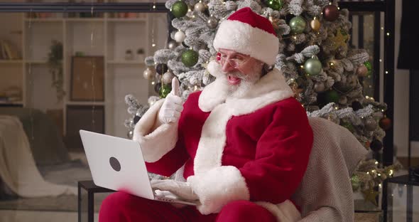Cheerful Santa Claus in His Residence with a Christmas Tree in the Background Using a Laptop alt