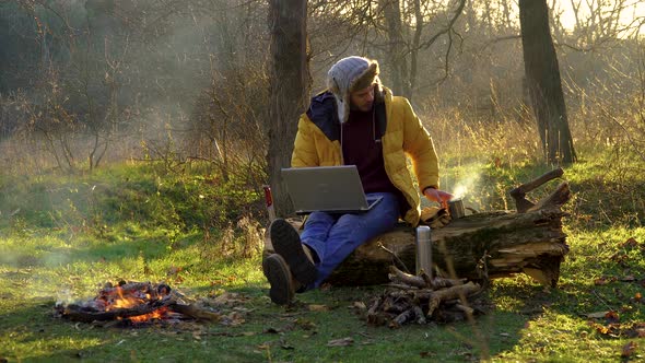 A Young Man Works at a Laptop Near the Fire in the Forest alt