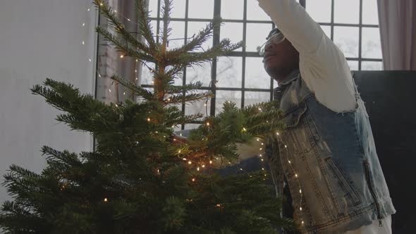 Focused Black Man in Glasses Putting Lights on Christmas Tree  Medium Closeup Shot alt