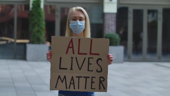 Young Woman Blonde in Medical Mask Stands with a Cardboard Poster ALL LIVES MATTER in a Public Place alt