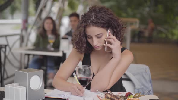 Beautiful Curly Girl Writing Down Information Sitting at the Table in Cafe Talking By Cellphone alt
