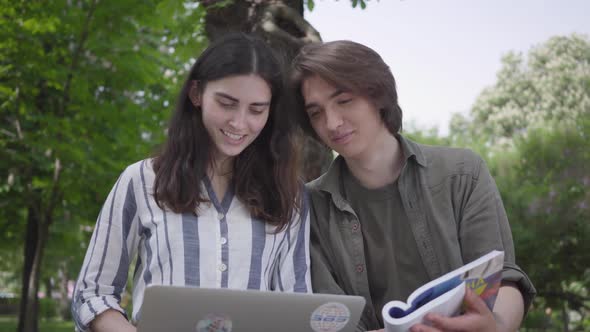 The Male and Female Students in Casual Clothes Sitting at the Bench, Girl Holding Laptop and Boy Has alt