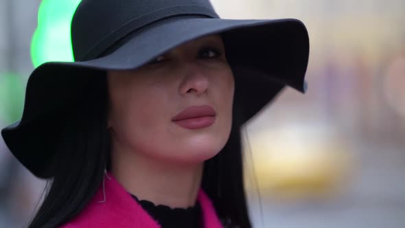 Portrait of a Brunette in a Black Hat and a Pink Coat Against a Background of Blurred Lights alt