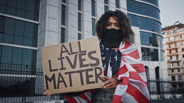 Darkskinned Protester Woman in Black Bandana on Face Wrapped in Flag of USA alt