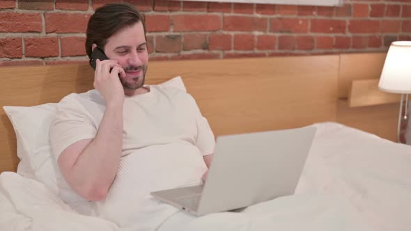 Casual Young Man with Laptop Talking on Smartphone in Bed alt