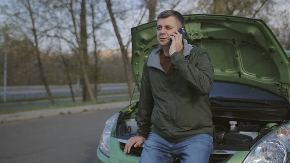 A Middleaged Man Crouched in Front of the Open Hood of the Car and Calls for Technical Assistance on