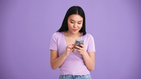 Cheerful Asian brunette woman wearing purple t-shirt texting by mobile alt