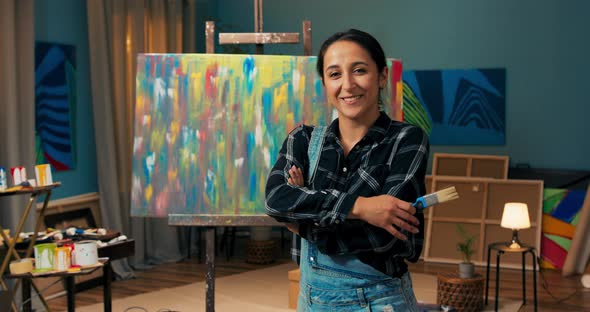 Female Wearing Denim Dungarees in a Hand Dirty From Paint Holds a Paint Brush Smile alt