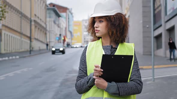 African American Female Engineer in Safety Vest Helmet Hardhat Engineer Standing in City Checking alt