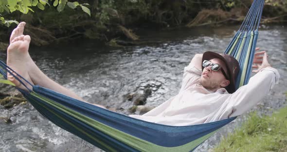 Handsome Man in a Hat and Sunglasses Lying and Swinging in a Hammock on a Summer Day
