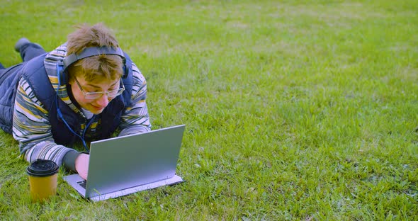 A Teenager in Headphones Lies on the Lawn in Front of a Laptop Monitor He Does His Homework or alt