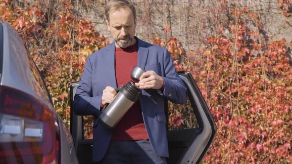 A Middleaged Handsome Caucasian Man Pulls a Vacuum Bottle Out of His Car and Drinks Tea in Fall