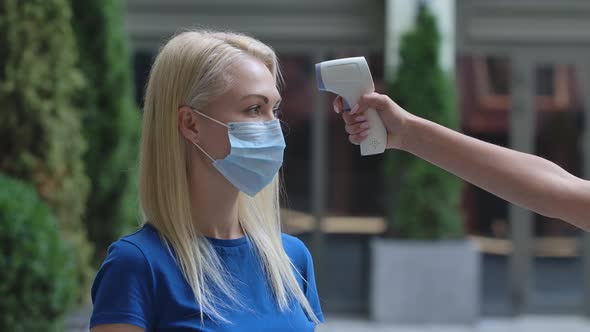 Portrait of a Charming Young Woman in a Protective Medical Mask Whose Temperature Is Measured with a alt