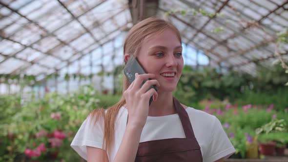 Young Girl Nursery Worker and Flower Greenhouse Speaks to a Client on the Phone