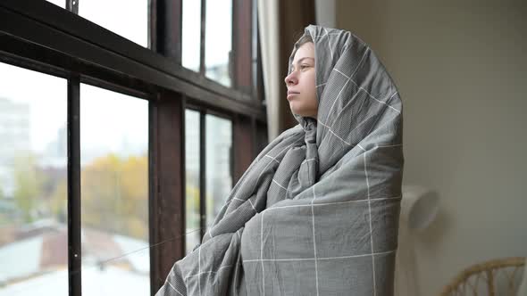 woman wrapped in a blanket stands near the window and looks out into the street alt