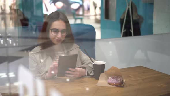 A Young Woman is Sitting Behind a Glass in a Cafe with a Tablet in Her Hands alt