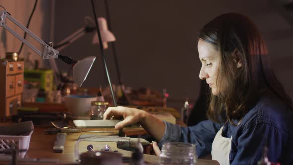 Profile of focused caucasian female jeweller sitting at desk, making jewelry in workshop alt