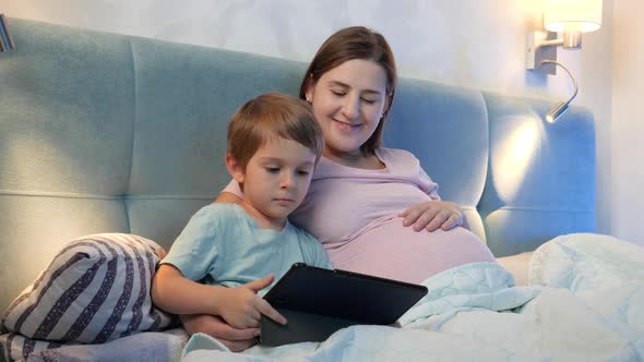 Happy Smiling Little Boy with Pregnant Mother Watching Cartoons on Tablet Computer in Bed at Night alt