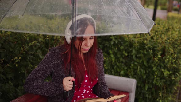 Young Beautiful Woman in Headphones with Transparent Umbrella Reading Book Sitting on Bench in City alt
