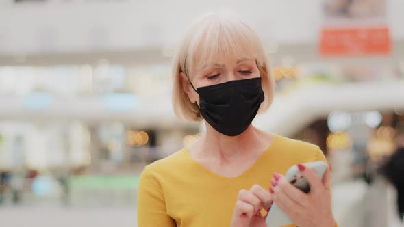 Serious Mature Middle Aged Woman in Medical Mask Walking Holding Smartphone Looking at Screen Typing alt