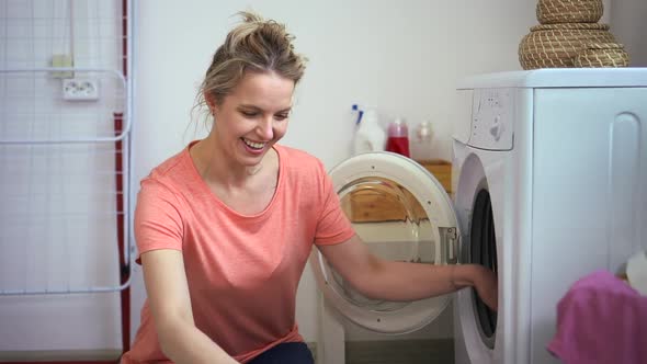 Woman Putting Clothes Into Washing Machine and Smiling While Sitting on Floor in Home Laundry Spbd alt