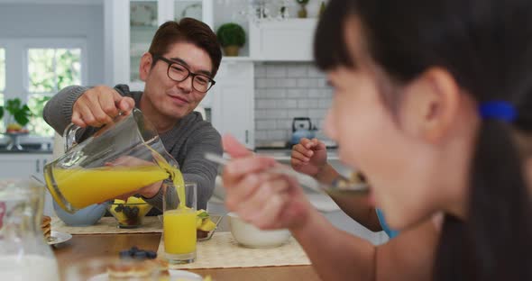 Happy asian father in kitchen having breakfast pouring juice for smiling son and daughter alt