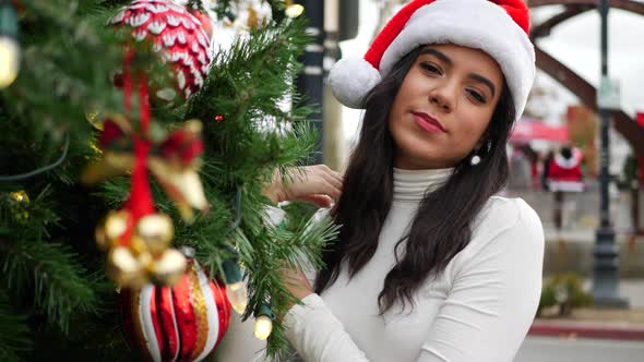A merry woman in a festive Santa Claus hat decorating a holiday Christmas tree with ornaments, jingl alt