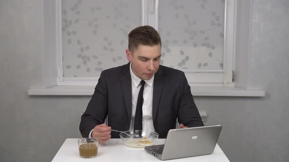 A Young Man in a Suit Eats Cereal and Drinks Coffee and Uses a Laptop alt
