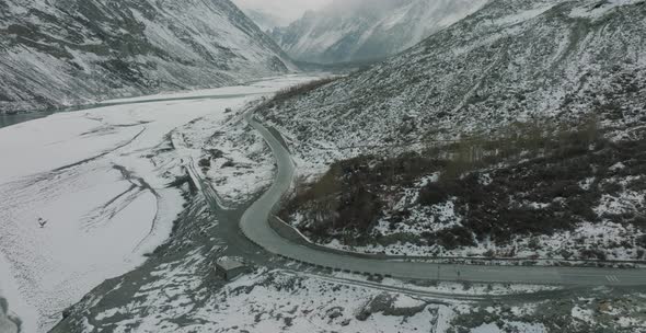 Aerial Flying Over Empty Road In Snow Covered Hunza Valley Landscape Beside Frozen River. Circle Dol alt