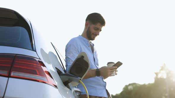 Bearded Man Standing Near an Electric Car and Making Time Adjustments on Smartphone