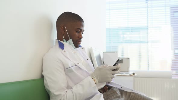 Doctor Sits in Medical Office During Free Time and Working on Smartphone