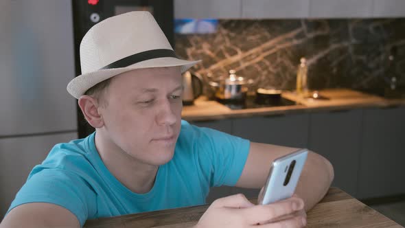 Man in Hat Sits at a High Wooden Table Using His Mobile Phone Kitchen Background alt